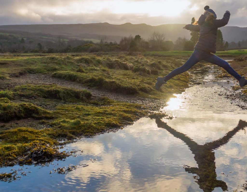 A woman jumps across a stream in the Highlands.
